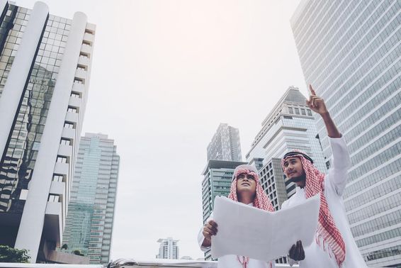Két arab tervező, kezükben egy tervrajzzal néznek a távolba, mögöttük felhőkarcolok láthatok.