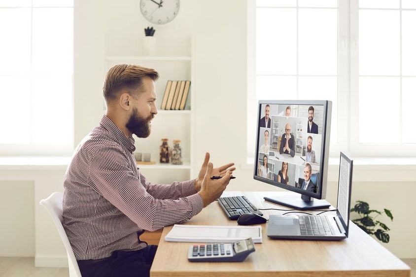 Egy ember ül az irodában az asztalon egy notebook, és egy számológép, elötte online webkamerás monitoron videokonferenciát folytat.