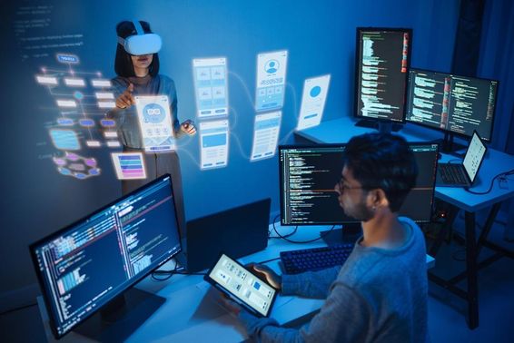 An app developer sits before a computer, holding a tablet, with two monitors displaying computer code.