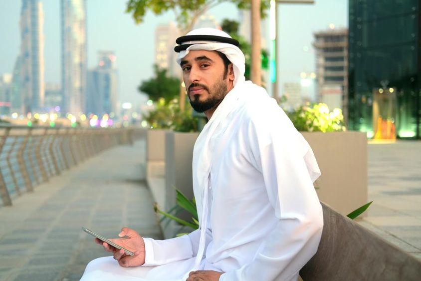 Emirati polgár ül egy padon, kezében egy telefon, háttérben a felhőkarcolok.