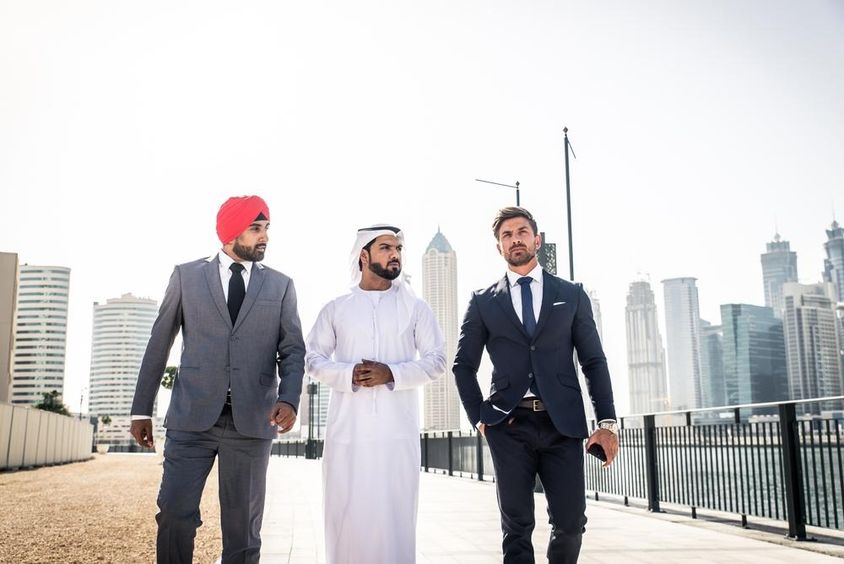 Ind, Emiráčan a Európan kráčajúci popri kanáli, mrakodrapy za nimi.