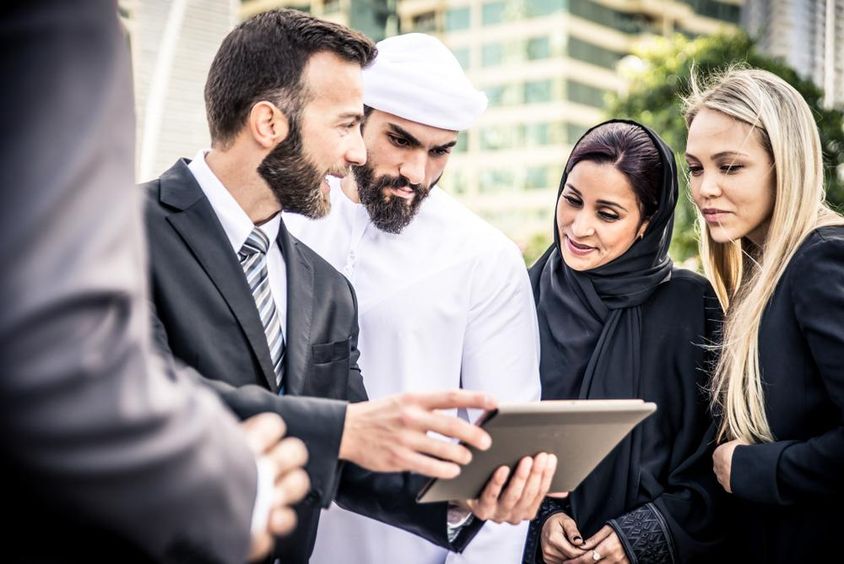 Egy európai és egy emriati férfi néz egy tabletet mellettük 2 nő egy szőke és egy emirati is figyeli. 