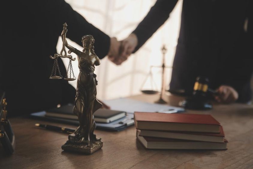 A lawyer is pictured negotiating with a client, with a Justitia statue visible on the table.