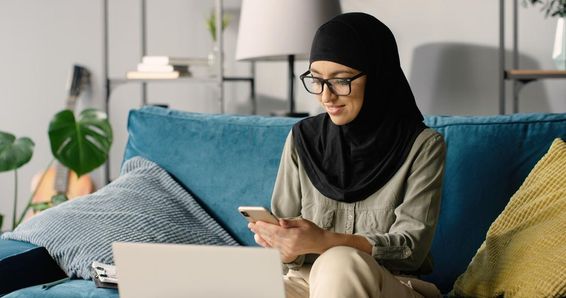 Arab lány hidzsábban dolgozik otthon okostelefon és laptop segítségével, miközben ül a kanapén.