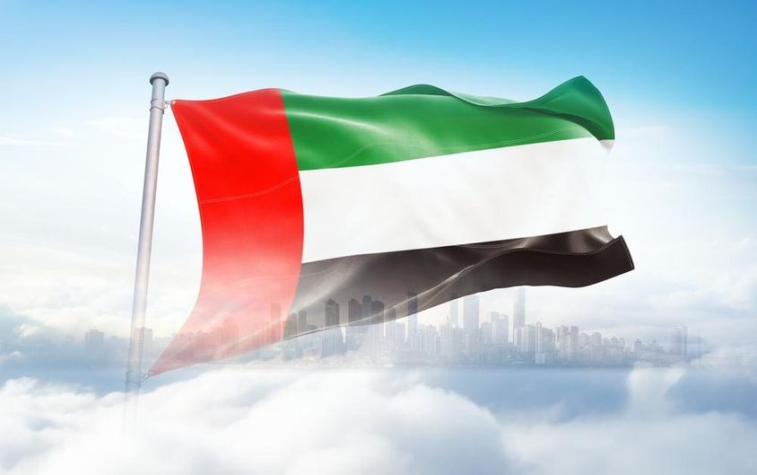 Bandeira dos EAU tremulando nas nuvens, arranha-céus ao fundo.