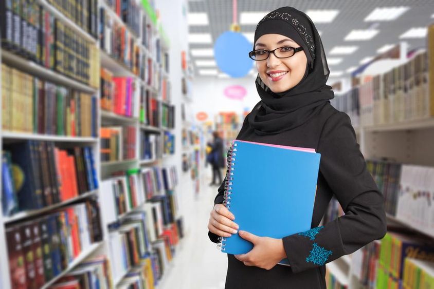 Arab student stands in the library with a notebook in hand.