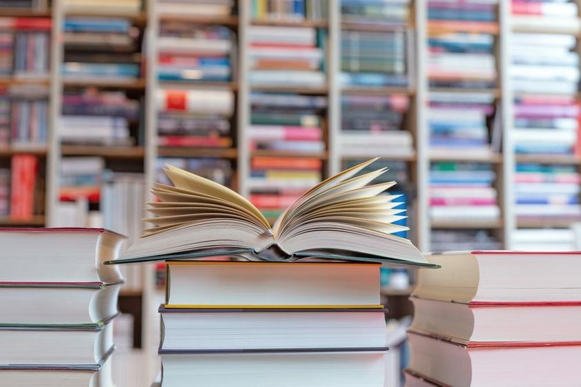 Open novel on top of a stack of books in front of a shelf.