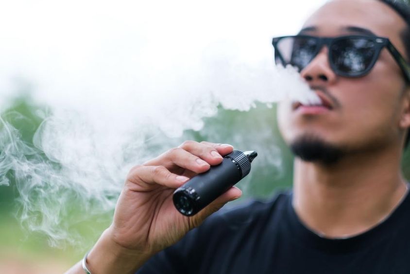 Asian man smoking with a vaping e-cigarette, exhaling smoke from his mouth and enjoying outdoors in summer.