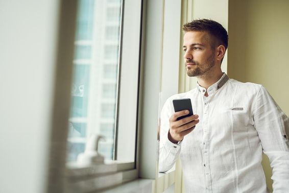 Üzletember állt az ablaknál, és tartja az okos telefonját.