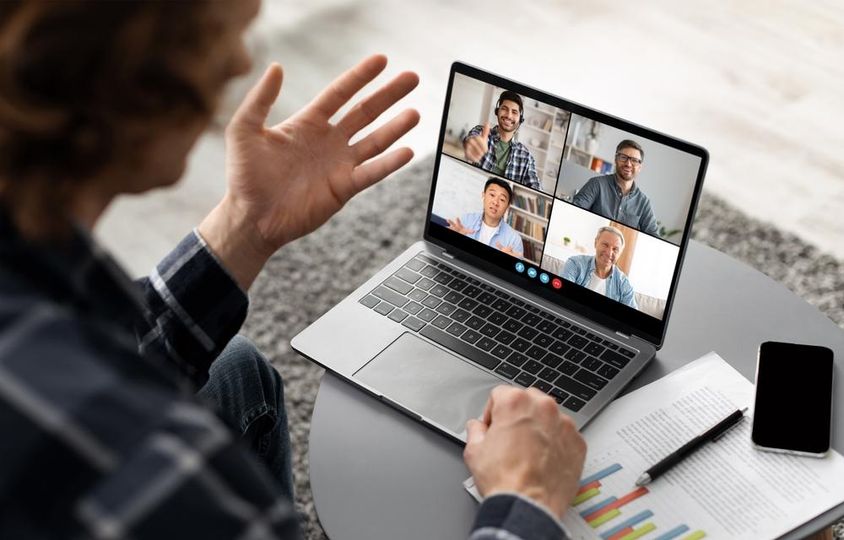 Egy férfi ül egy asztalnál, és egy videokonferencián vesz részt a laptopján.