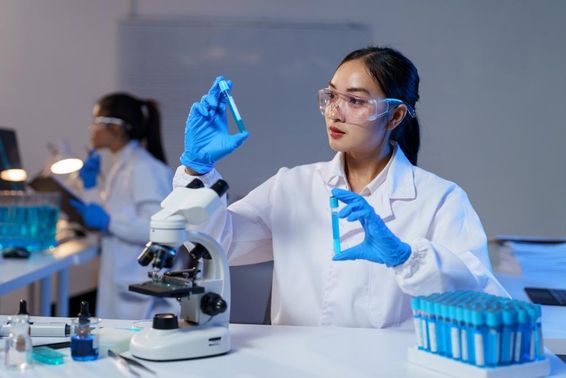 Asian female scientist in lab coat and gloves comparing two test tubes.
