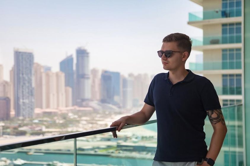 Egy fiatalember napszemüvegben, felhőkarcoló erkélyén áll, kilátással Dubaira.