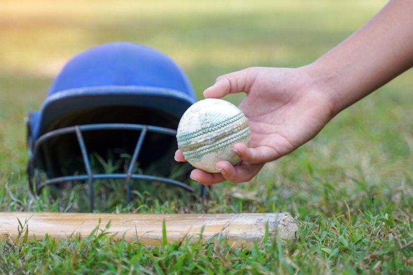 A player's hand picking up a cricket ball from among other cricket gear lying on the grass.