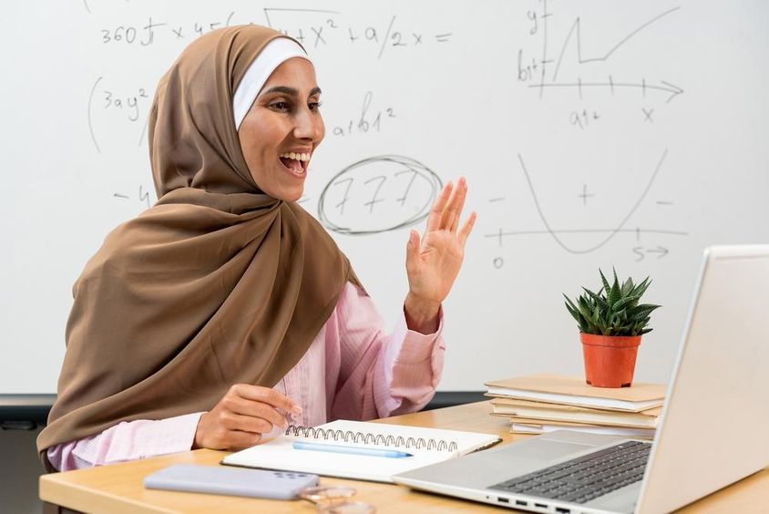 Női tanár hidzsábban integetve üdvözli a diákokat online óra alatt.