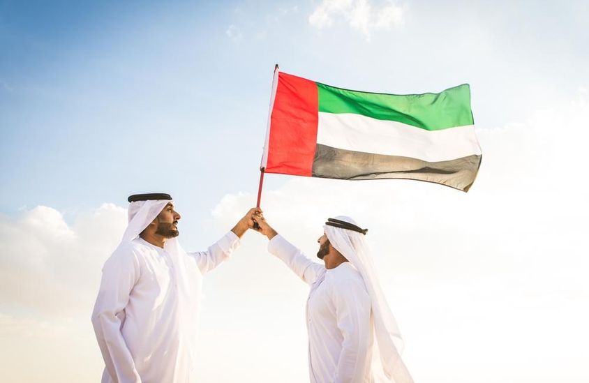 Arab férfiak, akik kandorát viselnek, sétálnak a sivatagban, kezükbe zászló.