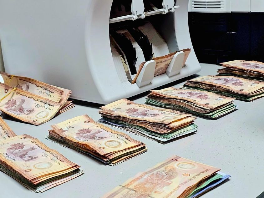 bankjegyszámláló gép, asztalán szépen elrendezett emirátusi dirham bankjegykötegekkel.