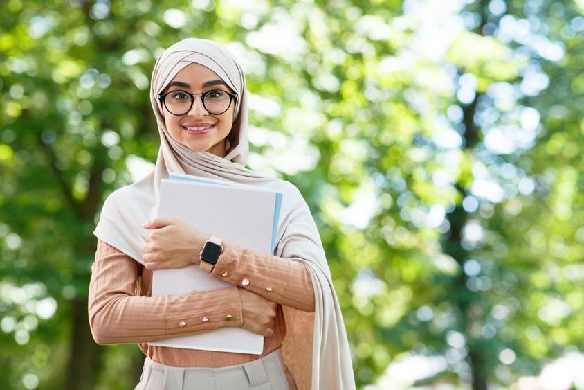 Vissza az iskolába, arab nő hidzsábban és szemüvegben könyveket tart a parkban.