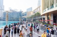 Turisták és helyiek sokszínű tömege sétál a Dubai Mall előtti forgalmas vízparti sétányon.